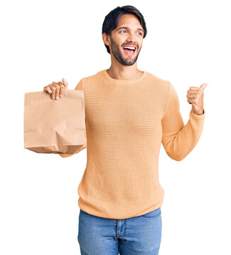 Handsome hispanic man holding take away paper bag pointing thumb up to the side smiling happy with open mouth