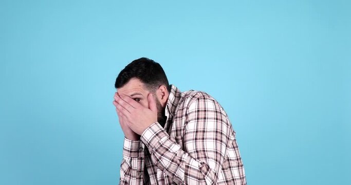Shocked bearded man, covers face with hands, look from fingers, isolated on blue studio background, free space. Problems, stress, fear emotions, scared film, facial expression. 