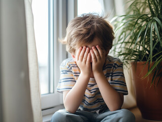 Portrait of adorable little boy sitting on the windowsill and crying. Upset child covering his face at home