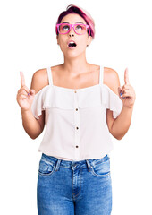 Obraz na płótnie Canvas Young beautiful woman with pink hair wearing casual clothes and glasses amazed and surprised looking up and pointing with fingers and raised arms.