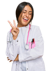 Beautiful hispanic woman wearing doctor uniform and stethoscope smiling with happy face winking at the camera doing victory sign. number two.