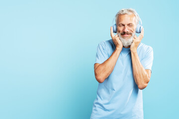 Handsome happy mature man, gray haired bearded hipster listening music wearing wireless headphones isolated on blue background, copy space. Technology, advertisement  concept