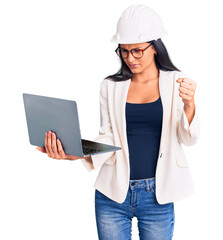 Young beautiful latin girl wearing architect hardhat holding laptop annoyed and frustrated shouting with anger, yelling crazy with anger and hand raised
