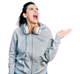 Young hispanic woman wearing gym clothes, using headphones and smart watch angry and mad screaming...