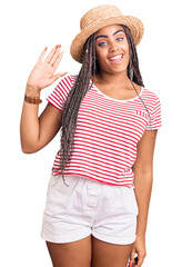 Young african american woman with braids wearing summer hat waiving saying hello happy and smiling, friendly welcome gesture