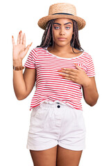 Young african american woman with braids wearing summer hat swearing with hand on chest and open...
