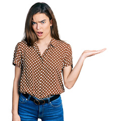 Young caucasian girl presenting with open palms, holding something in shock face, looking skeptical...