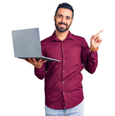 Young hispanic man holding laptop smiling happy pointing with hand and finger to the side