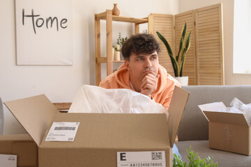 Thoughtful young man opening parcel at home - obrazy, fototapety, plakaty