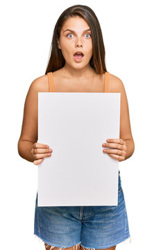 Young caucasian woman holding blank empty banner scared and amazed with open mouth for surprise, disbelief face