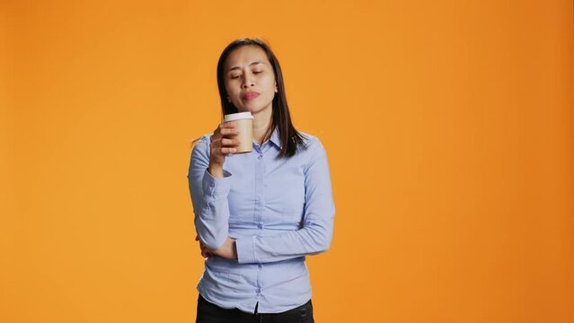 Filipino person savouring cup of coffee in studio, drinking caffeine refreshment for energy through the day. Young woman enjoying hot beverage while standing over orange background.