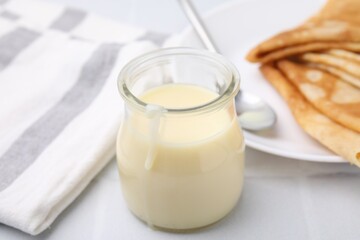 Obraz na płótnie Canvas Tasty condensed milk in jar and crepes on white table, closeup