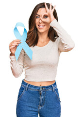 Young brunette woman holding blue ribbon smiling happy doing ok sign with hand on eye looking through fingers