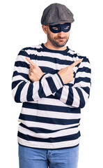Young handsome man wearing burglar mask pointing to both sides with fingers, different direction disagree