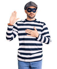 Young handsome man wearing burglar mask swearing with hand on chest and open palm, making a loyalty...