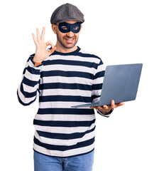 Young handsome man wearing burglar mask using laptop doing ok sign with fingers, smiling friendly gesturing excellent symbol
