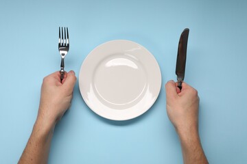 Man holding fork and knife near empty plate at light blue table, top view