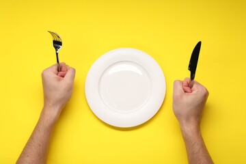 Man holding fork and knife near empty plate at yellow table, top view