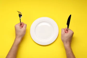 Fotobehang Man holding fork and knife near empty plate at yellow table, top view © New Africa