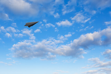 Black paper plane flying in blue sky with clouds