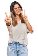 Young woman wearing casual clothes and glasses smiling looking to the camera showing fingers doing...