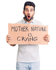 Young handsome man with beard holding mother nature is crying cardboard banner scared and amazed with open mouth for surprise, disbelief face