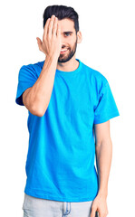 Young handsome man with beard wearing casual t-shirt covering one eye with hand, confident smile on face and surprise emotion.