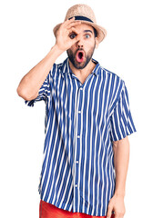 Young handsome man with beard wearing summer hat and striped shirt doing ok gesture shocked with surprised face, eye looking through fingers. unbelieving expression.