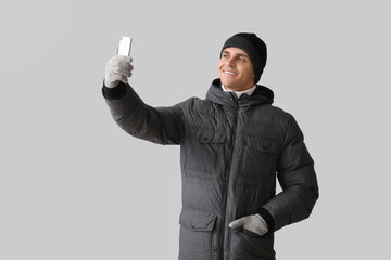 Young man in puffer jacket taking selfie on light background