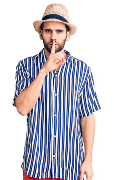 Young handsome man with beard wearing summer hat and striped shirt asking to be quiet with finger on lips. silence and secret concept.