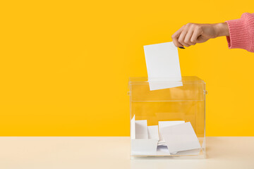 Hand putting voting paper in ballot box on yellow background - obrazy, fototapety, plakaty