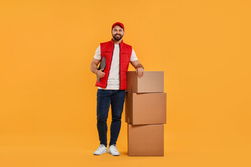 Happy young courier with clipboard and stack of parcels on orange background