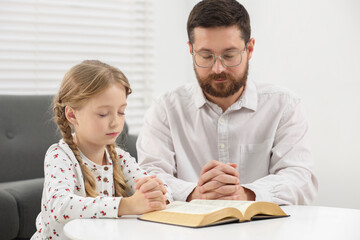 Girl and her godparent praying over Bible together at table indoors