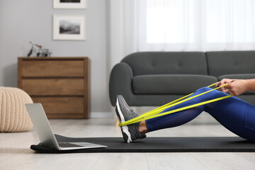 Woman doing exercise with fitness elastic band near laptop on mat at home, closeup