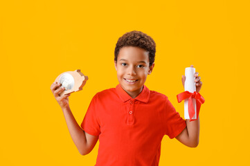 Little African-American boy with piggy bank and diploma on yellow background
