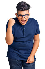 Little boy kid wearing casual clothes and glasses angry and mad raising fist frustrated and furious while shouting with anger. rage and aggressive concept.