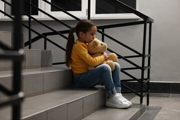 Child abuse. Upset little girl with teddy bear sitting on stairs indoors