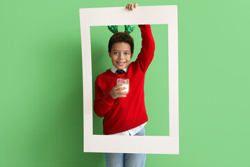 Little African-American boy in reindeer horns with glass cup of milk and frame on green background