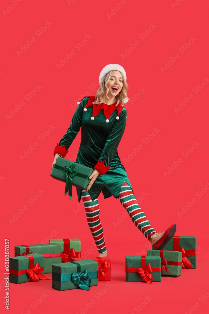 Wall mural happy young woman dressed as elf with christmas gifts on red background