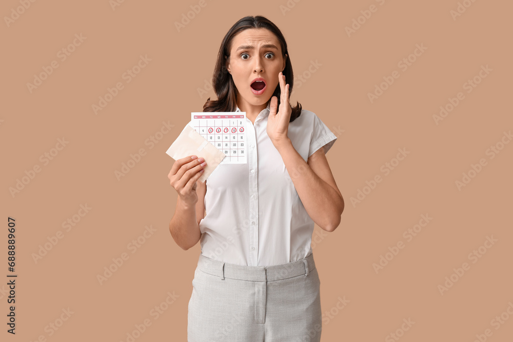 Wall mural beautiful shocked woman holding pad and calendar with marked days of menstruation on brown backgroun