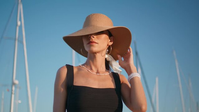 Elegant hat model posing in front yachts masts at summer embankment close up. 