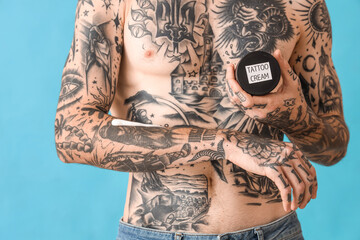 Tattooed man holding jar of tattoo cream on blue background, closeup