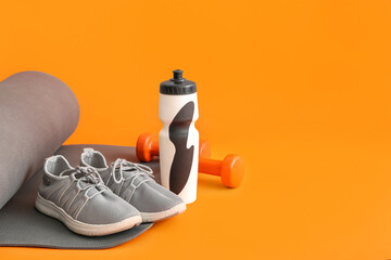 Yoga mat with sports bottle, dumbbells and sneakers on orange background
