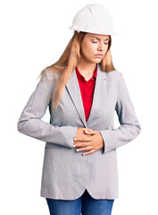 Beautiful young woman wearing architect hardhat with hand on stomach because indigestion, painful illness feeling unwell. ache concept.