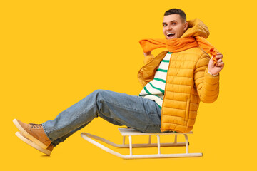 Young man in stylish puffer jacket with sled on yellow background
