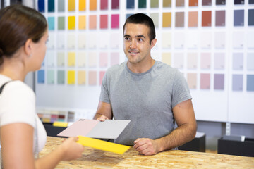 Portrait of positive polite salesman of painting supply store consulting young female client,...