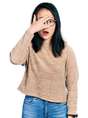 Fototapeta premium Young chinese woman wearing casual sweater and glasses peeking in shock covering face and eyes with hand, looking through fingers with embarrassed expression.