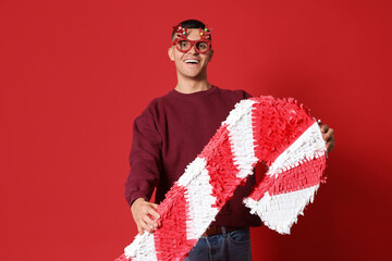 Happy young man in Christmas eyeglasses with candy cane pinata on red background