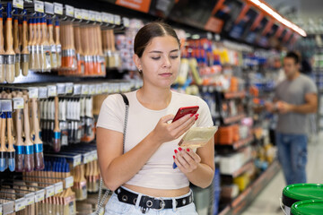 Positive young female customer scanning barcode on paintbrush with smartphone while choosing painting supplies in construction hypermarket, paying for item using mobile app..