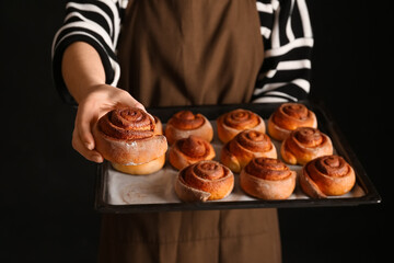 Woman holding baking tray with tasty cinnamon rolls on black background